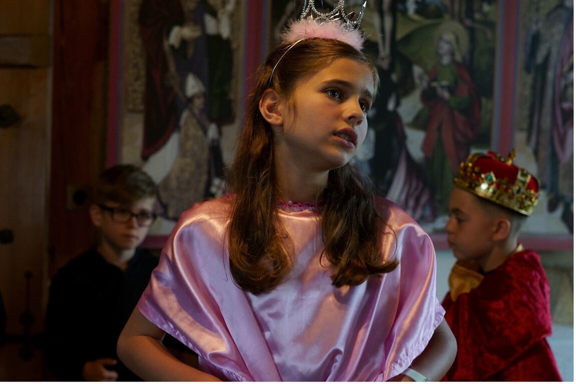 Ein junges Mädchen mit schillernd rosa Kleid und glitzerndem Krönchen mit rosa Federn am Ansatz in. Ihr Gesicht ist leicht zur Seite geneigt und zeigt einen konzentrierten leicht dramatischen Ausdruck. Hinter ihr etwas verschwommen stehen zwei kleine Jungen, der eine als König mit rotem Mantel und Krone verkleidet und der andere im schwarzen Hemd mit schwarzer Brille.