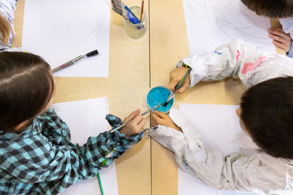 Vue d'en haut d'une table avec quatre enfants devant des papiers blancs avec des pinceaux et de la couleur bleu.