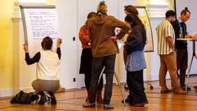 Mehrere Personen stehen um Stehtische herum und lesen oder schreiben. Eine Frau kniet links vorne vor einem Flipchart und schreibt mit blauem Filzstift eine Bullet-Point-Liste mit Notizen. 