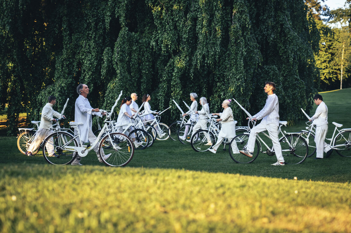 Auf einer Wiese im Park mit einem grossen grünen Baum mit bis zum Boden hängenden Ästen stehen sich in zwei Reihen weiss gekleidete Menschen gegenüber. Jede Person schiebt ein weisses Velo auf dem an der Lenkstange das Horn eines Einhorns befestigt ist.