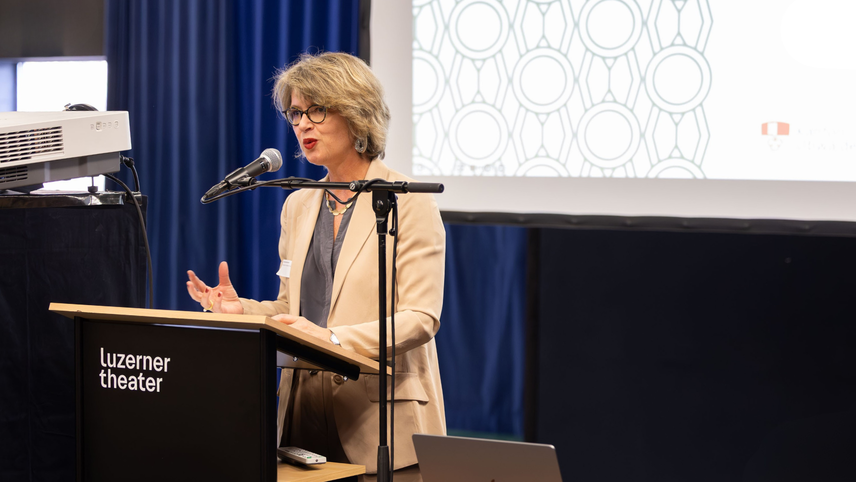 Eine Frau mit dunkler Hornbrille und rotem Lippenstift, schulterlangem blondem Haar, in einem beigen Blazer spricht ins Mikrofon an einem Rednerpult.