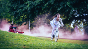 Sur un fond d'arbres et de poussière rose qui monte du sol, une femme à vélo vêtue d'un tutu blanc dévale une pente herbeuse. Sur sa gauche, en haut de la pente, une figure vêtue de pourpre la suit, assise sur une luge.