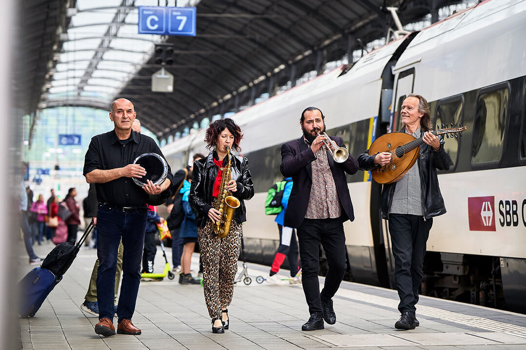 Christian Fotsch mit Ssassa vier Musiker_Innen gehen nebeneinander auf dem Perron eines Bahnhofs musizierend entlang mit Trommel, Saxophon, Trompete und Oud.