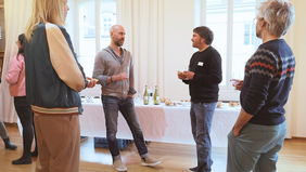 In einem hellen Raum mit grossen Fenstern und weissen vorhängen stehen Menschen vor einem Buffet mit Essen und Getränken auf weisser Tischdecke. In der Mitte stehen sich zwei Männer im Gespräch gegenüber. Der eine mit kurzem braunen Haar und silbrigem Ohrring, der andere mit glatt geschorenem Kopf und blondem Bart. Sie halten Weingläser in der Hand.