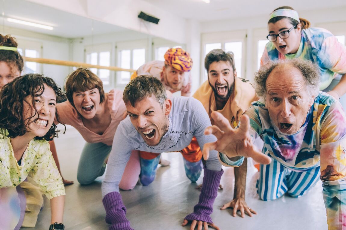 Auf dem Foto sind sieben Personen, die auf allen Vieren auf die Kamera zukriechen und mit offenen Mündern und gefletschten Zähnen Grimassen schneiden. Sie tragen bunte gemusterte Kleider. Im Hintergrund ist die Spiegelwand und die Ballettstange eines Tanzstudios erkennbar.