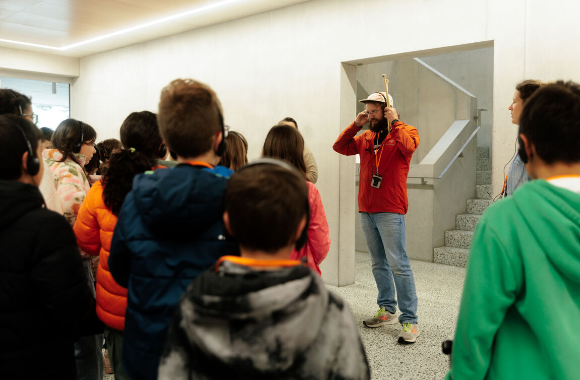 Un groupe d'enfants portant des écouteurs face à un homme barbue avec casquette avec des baguettes de batterie dans la main qui lui aussi met des écouteurs sur sa tête.