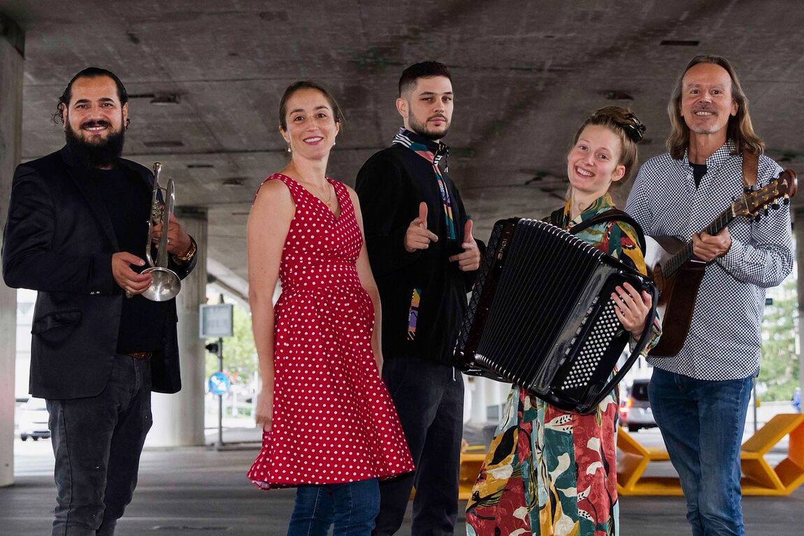 Eine Gruppe Musiker_Innen in einem überdachten Parkplatz lacht in die Kamera: Drei Männer und zwei Frauen mit Trompete, Akkordeon und Oud. 